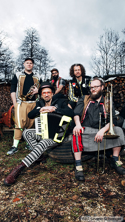 Die fünf Musiker der Band Kellerkommando posieren im Freien vor einem Holzstapel. Sie tragen auffällige, bunte Kleidung und halten ihre Instrumente, darunter ein Akkordeon, eine Trompete und eine Posaune.